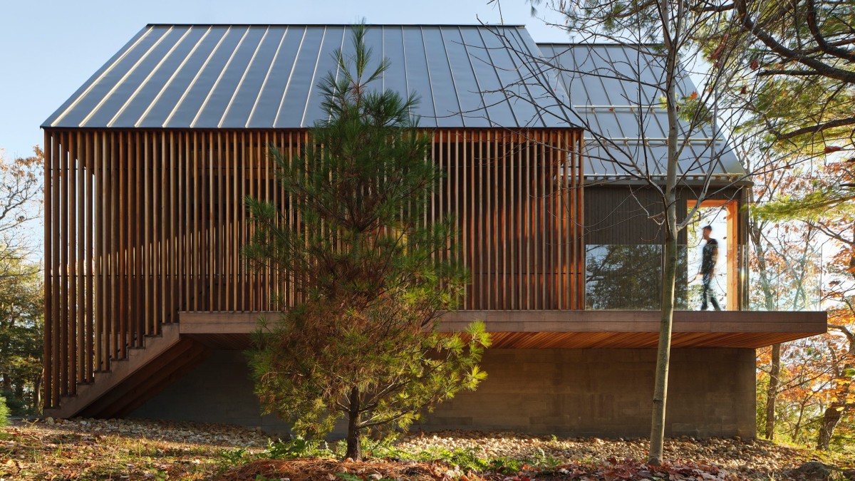 El acceso a la cabaña se realiza a través de una entrada lateral enmarcada por una pantalla de cedro, que añade calidez y contraste con el resto del revestimiento exterior.