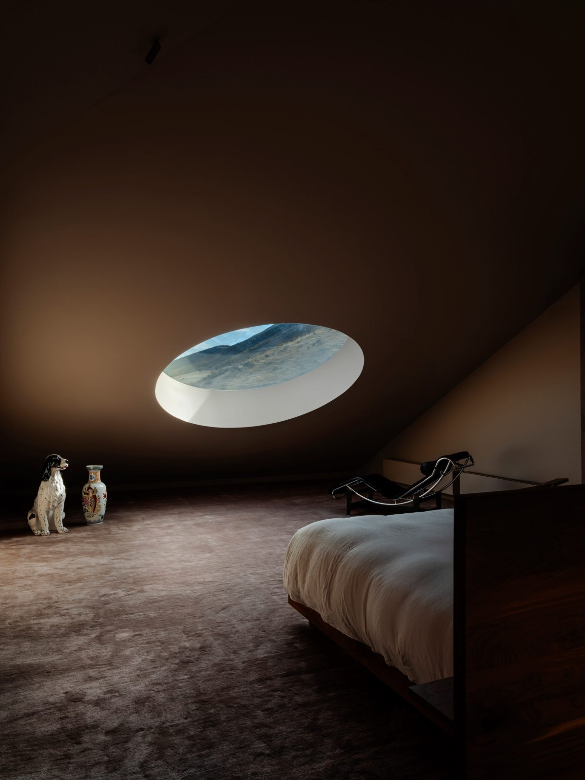 El altillo en buhardilla alberga un segundo dormitorio donde disfrutan de una claraboya circular que, situada a baja cota, enmarca las montañas lejanas.